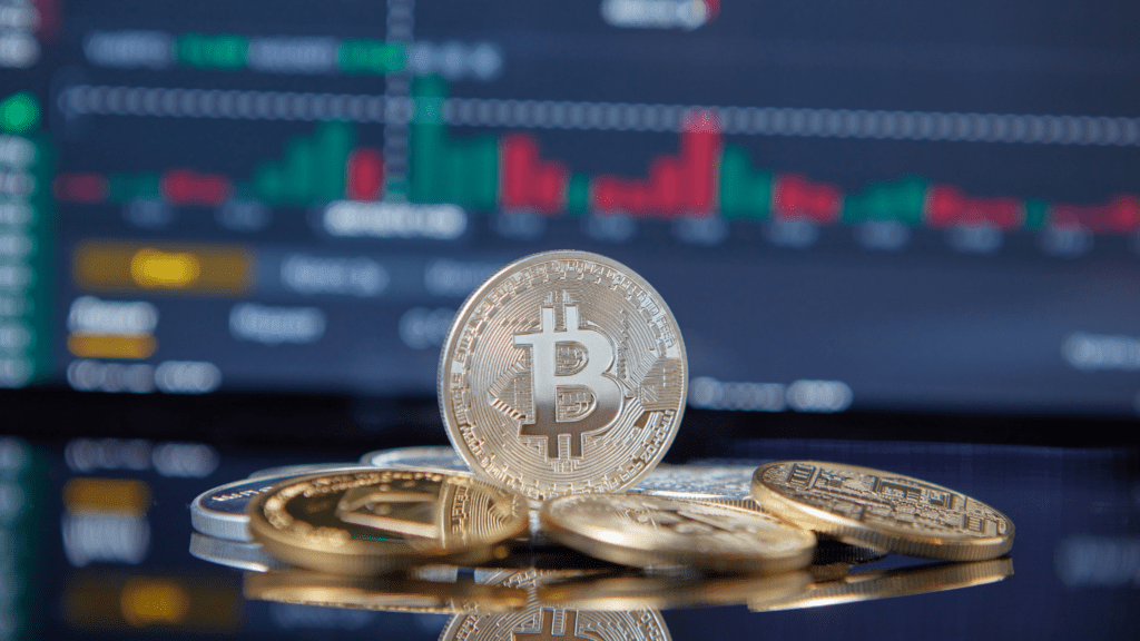 Bitcoin and gold coins on a table with a stock market screen in the background